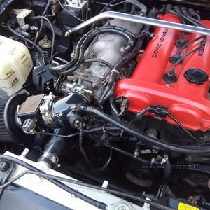 May include: Close-up of a black engine with a red valve cover. The valve cover has the text "DOHC 16-VALVE" and "mazda" printed on it. The engine has a black air filter and a black intake manifold.