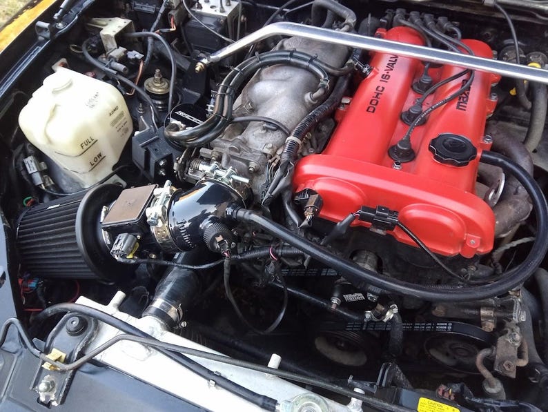 May include: Close-up of a black engine with a red valve cover. The valve cover has the text "DOHC 16-VALVE" and "mazda" printed on it. The engine has a black air filter and a black intake manifold.