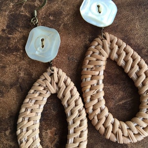May include: A pair of woven rattan earrings with oval shapes and white button accents. The earrings have a natural brown color and are made of woven rattan.