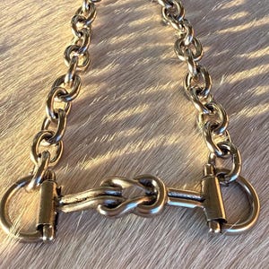 May include: A silver-toned horse bit with a chain, designed for equestrian use. The bit features a central knot design and rounded rings on each end. The chain is composed of oval links.
