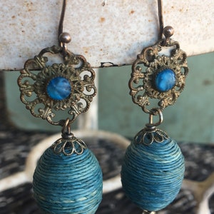 May include: A pair of dangle earrings with blue glass stones set in ornate gold-toned metal filigree. The earrings feature a blue woven bead hanging from each filigree.