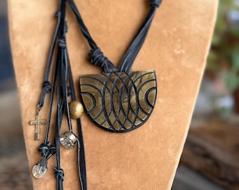 May include: A black leather cord necklace featuring a large, semi-circular, olive-green pendant with a carved geometric pattern. The necklace includes multiple dangling charms, including a small metal cross, a gold-toned bead, and a coin-style pendant.