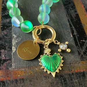 May include: A beaded necklace with iridescent green beads. The necklace features a gold-colored clasp and charms, including a heart-shaped pendant, a small cross with a gem, and a round charm with the word "Love" engraved on it.
