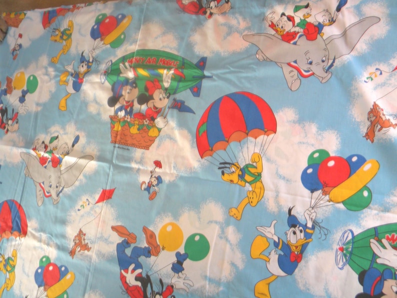 May include: Light blue fabric featuring a repeating pattern of Disney characters in the sky. Mickey and Minnie Mouse are in a basket, Pluto is parachuting, and Donald Duck is holding balloons. Dumbo and other characters are also present.