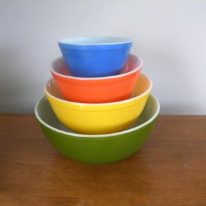 May include: A stack of four colorful mixing bowls. The bowls are nested, with the largest green bowl at the bottom, followed by yellow, orange, and blue bowls. Each bowl has a white rim.