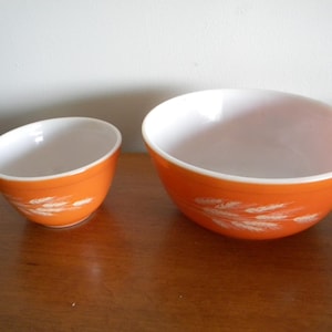 May include: Two orange mixing bowls with white interiors. The bowls have a wheat stalk design in white. The bowls are sitting on a wooden surface.