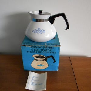 May include: A white CorningWare 6-cup teapot with a black handle and lid, decorated with blue floral patterns. The teapot is on a blue box with the text "6 CUP TEAPOT" and a drawing of the teapot. A small instruction sheet is in front.