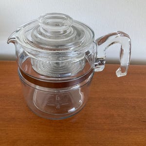 May include: A clear glass coffee pot with a lid, handle, and internal filter. The pot has a brown band around the middle and measurement markings. The handle is curved and attached to the side of the pot.