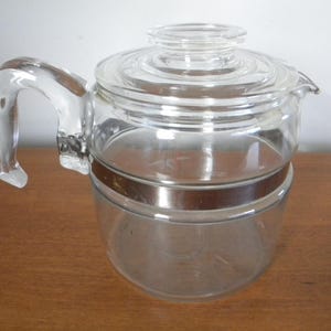 Peut inclure: Cafetière en verre transparent avec une anse et un couvercle. La cafetière est ornée d'une bande argentée au milieu. La poignée est incurvée et fixée sur le côté. Posée sur une surface en bois.