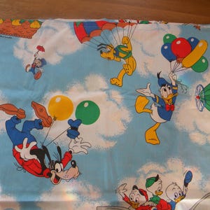 May include: Light blue fabric featuring cartoon characters like Mickey Mouse, Donald Duck, and Goofy. The design includes images of balloons, parachutes, and a hot air balloon, set against a sky with clouds. The fabric is likely for children's bedding.