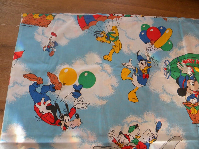May include: Light blue fabric featuring cartoon characters like Mickey Mouse, Donald Duck, and Goofy. The design includes images of balloons, parachutes, and a hot air balloon, set against a sky with clouds. The fabric is likely for children's bedding.
