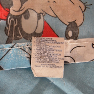 May include: Close-up of a light blue fabric with a cartoon character design in red, black, and gray. A tag with washing instructions is attached. The tag reads "Made in U.S.A."