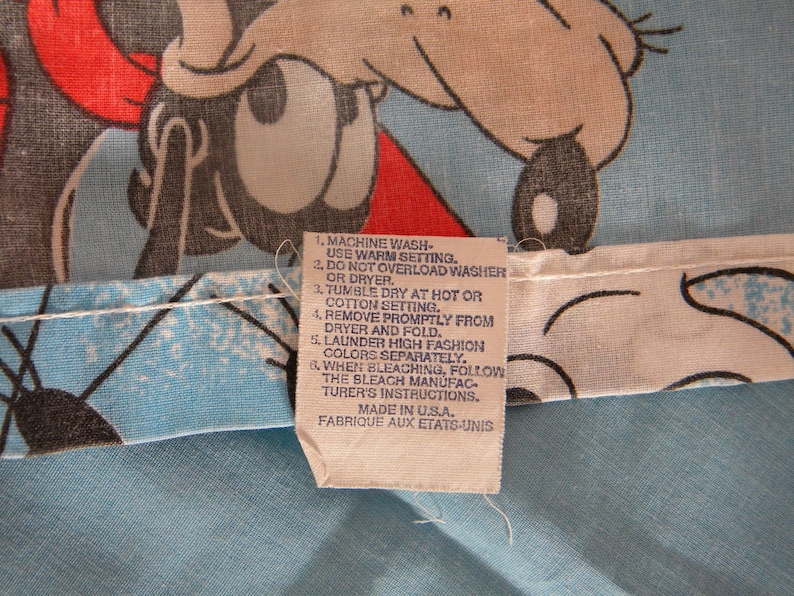 May include: Close-up of a light blue fabric with a cartoon character design in red, black, and gray. A tag with washing instructions is attached. The tag reads "Made in U.S.A."