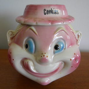 May include: Vintage ceramic cookie jar shaped like a smiling clown face. The jar is primarily pink and white, with blue eyes and a hat that reads "Cookies". The clown has a large, expressive smile.