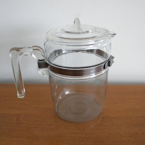 May include: A clear glass coffee pot with a metal band around the middle and a glass lid. The pot has a handle and a spout.
