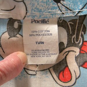 May include: Close-up of a fabric tag from Pacific Home Products. The tag reads "50% Cotton, 50% Polyester, Twin." The tag is on a light blue fabric with a cartoon character design. The tag also includes the address 285 Avenue of the Americas, New York, NY 10019.