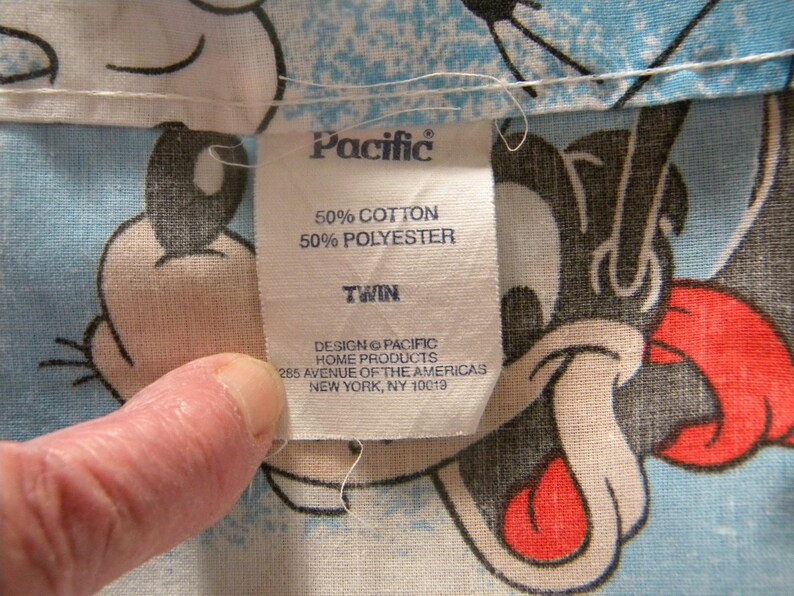May include: Close-up of a fabric tag from Pacific Home Products. The tag reads "50% Cotton, 50% Polyester, Twin." The tag is on a light blue fabric with a cartoon character design. The tag also includes the address 285 Avenue of the Americas, New York, NY 10019.