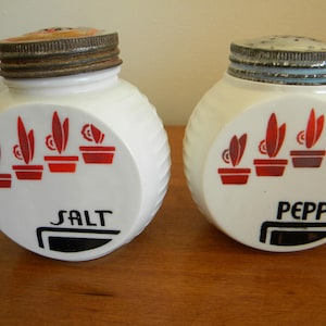 May include: White ceramic salt and pepper shakers with red and black designs. The shakers have a ribbed texture and feature a row of stylised plants in pots. The salt shaker says "SALT" and the pepper shaker says "PEPPER".