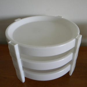 May include: A white, tiered serving tray with a round top and three curved tiers. The tray has vertical supports on the sides. The tray is made of a glossy, smooth material and sits on a wooden surface.