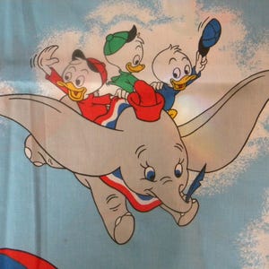 May include: A whimsical illustration of Dumbo the elephant soaring through a blue sky, carrying three cartoon duck characters. The ducks wear red, green, and blue outfits, and the elephant has a red and blue collar. A kite is visible in the background.