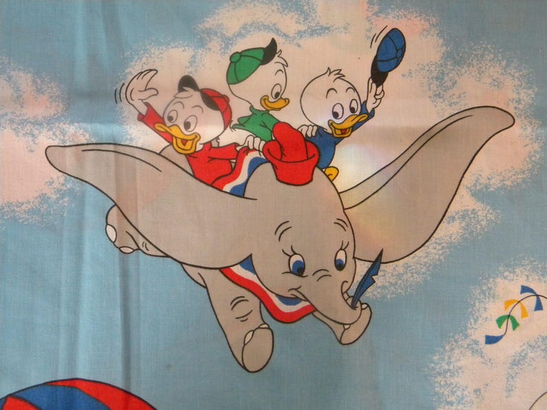 May include: A whimsical illustration of Dumbo the elephant soaring through a blue sky, carrying three cartoon duck characters. The ducks wear red, green, and blue outfits, and the elephant has a red and blue collar. A kite is visible in the background.
