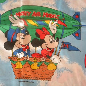 May include: A vintage fabric featuring Mickey and Minnie Mouse in a hot air balloon. Mickey wears a blue suit and Minnie a red dress. The green airship reads "Mickey Air Mobile". The background is a light blue sky with clouds and other balloons.