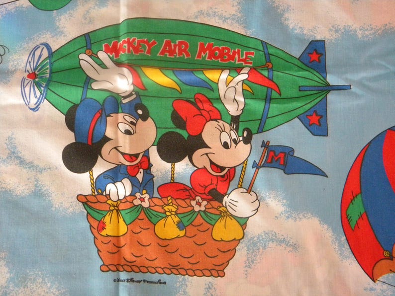 May include: A vintage fabric featuring Mickey and Minnie Mouse in a hot air balloon. Mickey wears a blue suit and Minnie a red dress. The green airship reads "Mickey Air Mobile". The background is a light blue sky with clouds and other balloons.
