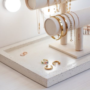 Puede incluir: Un expositor de joyas de cerámica blanca con cadenas, pulseras y anillos de oro. El expositor tiene una base rectangular y dos postes redondos. Las joyas están dispuestas de forma decorativa.