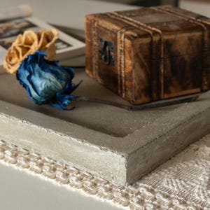Puede incluir: Una bandeja de hormigón gris con un borde decorativo se encuentra sobre una superficie de crochet blanco. Una caja de madera con un diseño tallado y un arreglo de flores secas con flores azules y amarillas están sobre la bandeja.