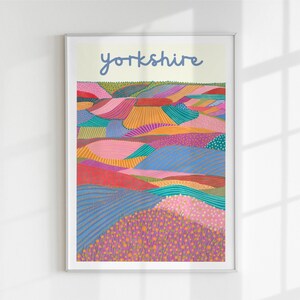 May include: A framed art print with a vibrant, abstract landscape design. The artwork shows rolling hills in shades of pink, blue, orange, and green, with the word "yorkshire" in blue script at the top. The print is displayed on a white wall.