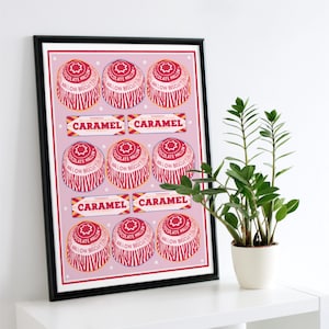 May include: A framed print with a repeating pattern of red and white chocolate marshmallow biscuits and caramel candy bars on a pink background. The print is in a black frame and displayed on a white shelf next to a potted plant.