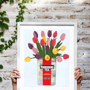 May include: A framed print of a still life featuring a bouquet of colourful tulips in a glass jar with a Yorkshire Tea label. The jar is filled with water and the tulips are arranged in a loose, natural way. The background is a light blue colour.