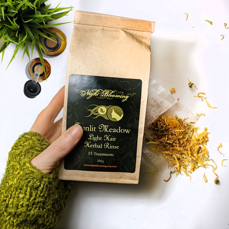 May include: A brown paper bag with a label that reads "Night Blooming" and "Sunlit Meadow Light Hair Herbal Rinse 15 Treatments 100g www.nightblooming.etsy.com". The bag contains a tea bag filled with dried yellow flower petals.