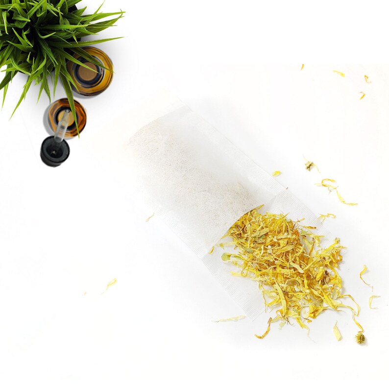 May include: A white tea bag filled with dried yellow flower petals. The tea bag is on a white surface with some petals scattered around it.