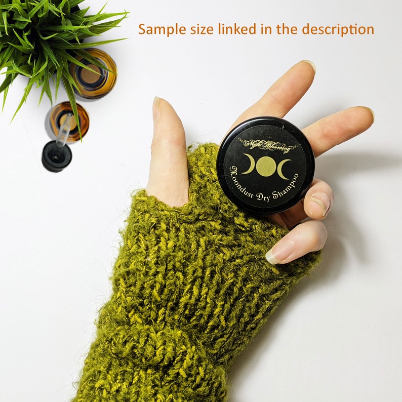 May include: A small black jar with a gold label that reads "Moondust Dry Shampoo" and a triple moon symbol. The jar is being held in a hand wearing a green knitted fingerless glove.
