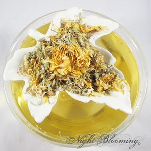 May include: A white butterfly-shaped tea infuser filled with dried herbs and flowers, including yellow calendula petals, in a clear glass bowl with a yellow liquid.