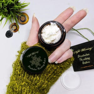 May include: A small jar of white salve with a black and gold label that reads "Night Blooming Feathalas Healing Salve". The jar is being held by a hand wearing a green knitted mitten.