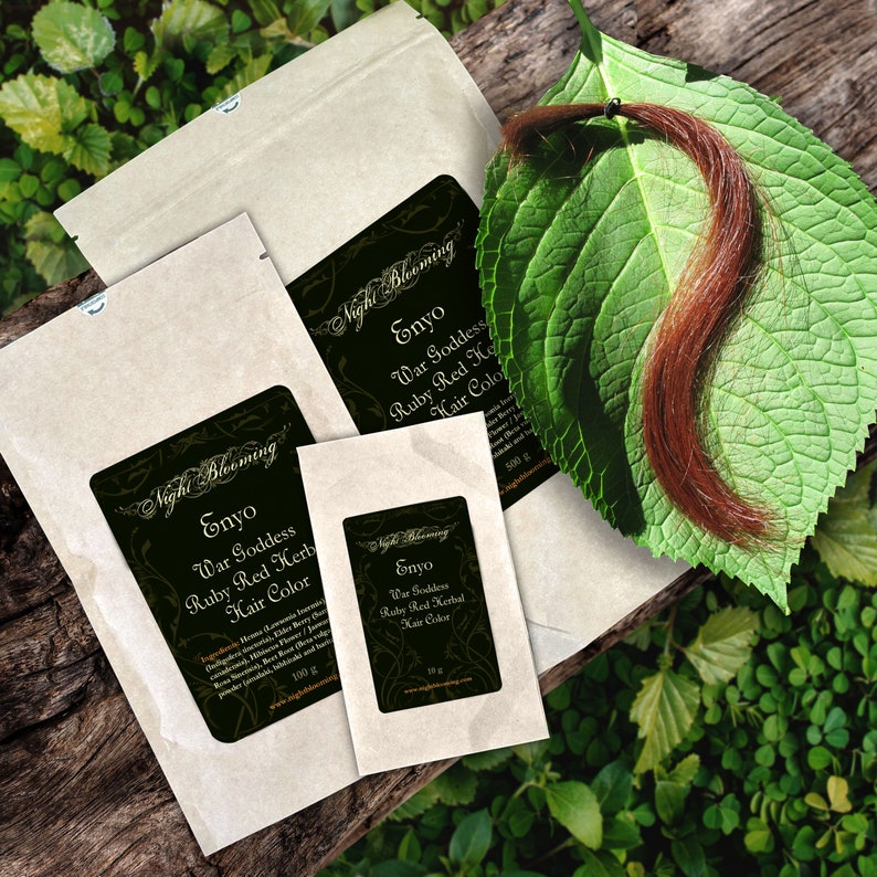 May include: Three brown paper packets with black labels that say "Night Blooming" and "Enyo War Goddess Ruby Red Herbal Hair Color" in different sizes. The packets are on a wooden surface with green leaves and a strand of red hair.