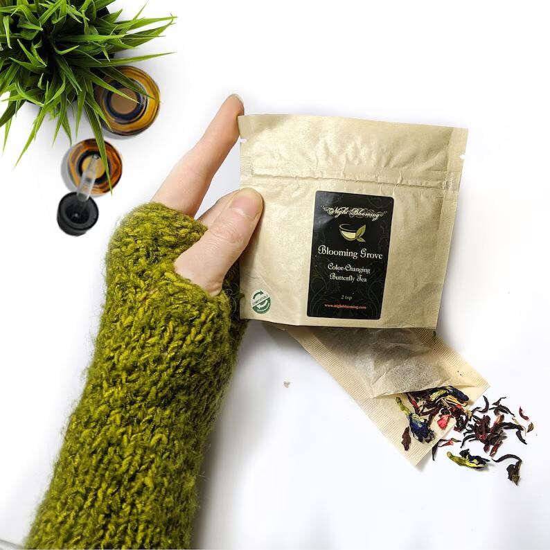 May include: A small brown paper bag with a label that reads "Blooming Grove Color Changing Butterfly Tea 2-cup". The bag is open and a tea bag is spilling out, revealing loose blue, red, and green tea leaves.