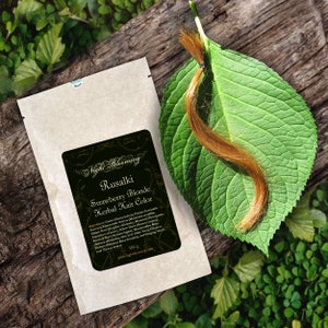 May include: A small brown paper packet with a label that reads "Night Blooming Rusalki Strawberry Blonde Herbal Hair Color". The label lists ingredients and a website address. A strand of light brown hair is on a green leaf.