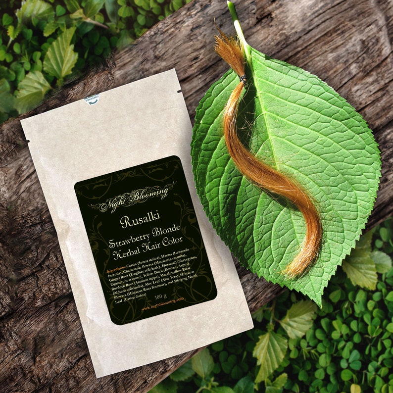 May include: A small brown paper packet with a label that reads "Night Blooming Rusalki Strawberry Blonde Herbal Hair Color". The label lists ingredients and a website address. A strand of light brown hair is on a green leaf.