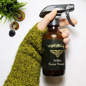 May include: A brown glass bottle with a black label that says "Night Blooming Selkie Herbal Detangler 8 oz". The bottle has a black spray nozzle and is being held by a hand wearing a green knitted mitten.