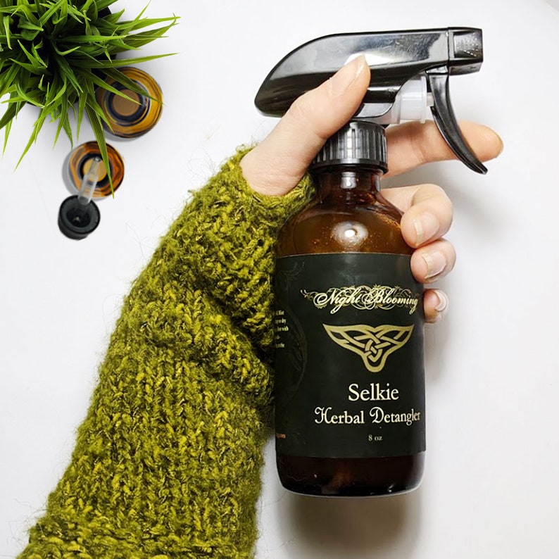 May include: A brown glass bottle with a black label that says "Night Blooming Selkie Herbal Detangler 8 oz". The bottle has a black spray nozzle and is being held by a hand wearing a green knitted mitten.