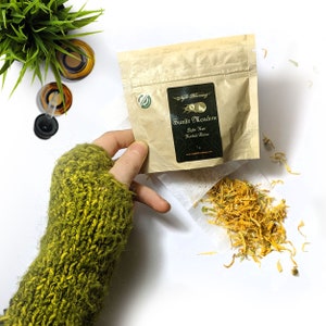 May include: A kraft paper packet labeled "Sunlit Meadow Light Rain Herbal Rinse" is held over a small pile of dried orange flower petals. The packet has a black label with white text and a green circular sticker. A green and yellow knitted sleeve is visible.