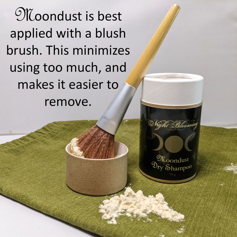 May include: A white container with a black and gold label that reads "Night Blooming Moondust Dry Shampoo 75g". A makeup brush with a wooden handle and a white and silver ferrule is resting on a small brown container filled with a light brown powder. The container and brush are on a green fabric surface.