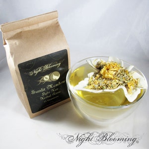 May include: A brown paper bag with a black label that reads "Night Blooming Sunlit Meadow Light Hair Herbal Rinse 15 Treatments". A glass bowl with a white ceramic flower-shaped infuser filled with yellow and green herbs is in the background.