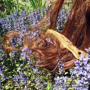 May include: A wooden comb with intricate carvings lies on a bed of purple flowers. The comb is brown with a light wood grain and has a long, narrow handle. The flowers are in bloom and have a delicate, feminine appearance.