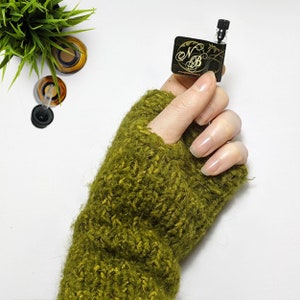 Puede incluir: Un peque&ntilde;o frasco de perfume de vidrio con una tapa negra y una etiqueta que dice "NB" en letras doradas. El frasco est&aacute; siendo sostenido por una mano que lleva un calientamanos de punto verde.