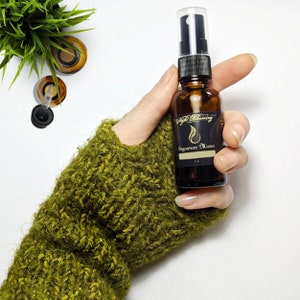 May include: A brown glass bottle with a black spray nozzle and a label that reads "Night Blooming Signature Mister". The bottle is being held in a hand wearing a green knitted mitten.