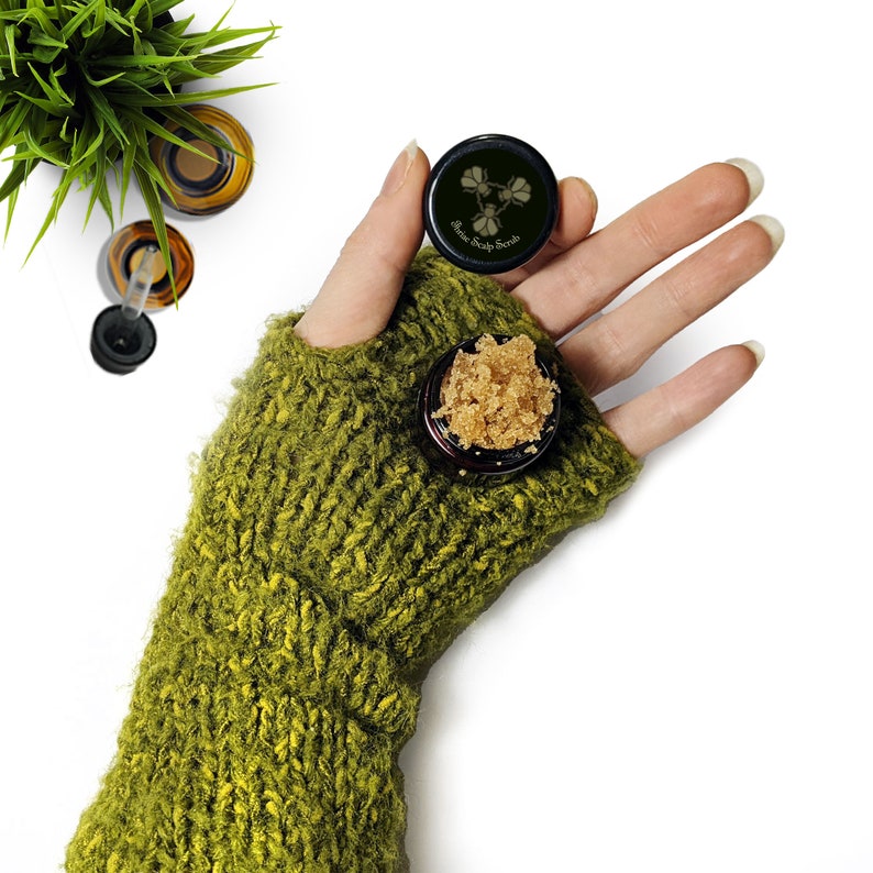 May include: A small jar of brown sugar scrub with a black lid. The lid has a white label with the text "Sugar Scalp Scrub". The jar is being held by a hand wearing a green knitted fingerless glove.